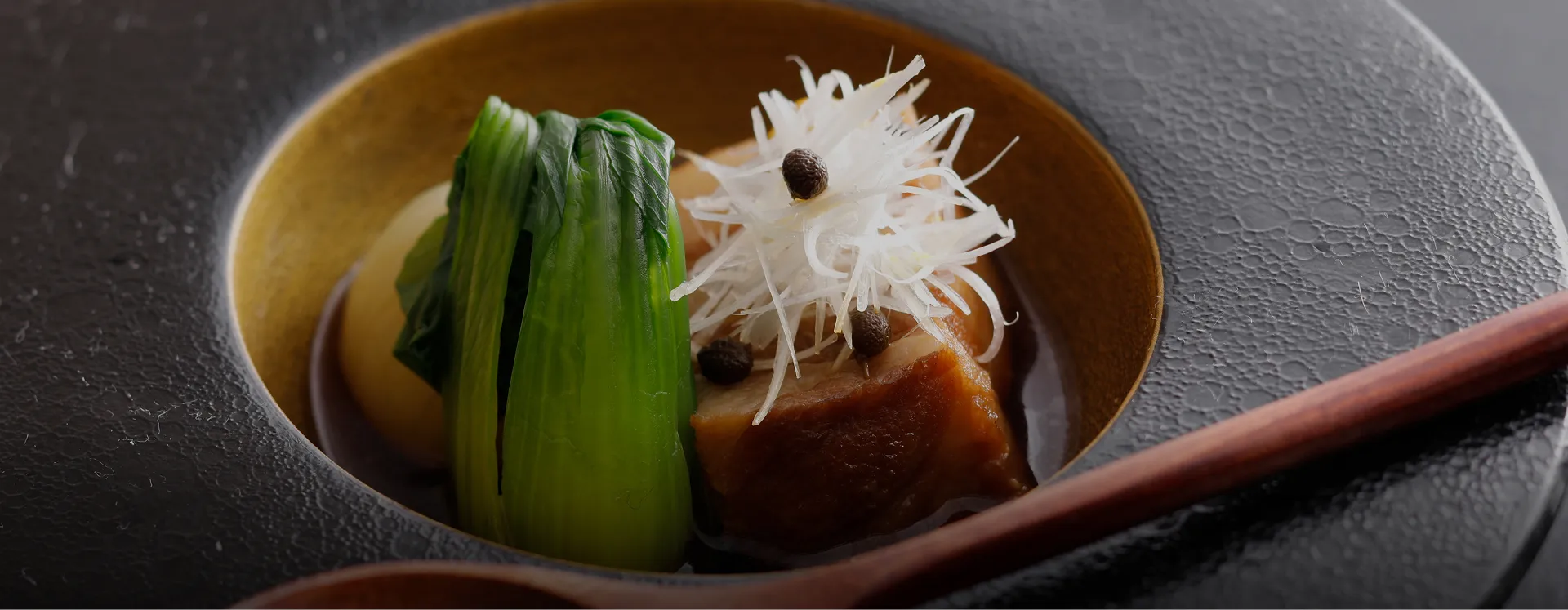 黒と金の器に盛り付けられた、豚の角煮と色鮮やかな野菜の煮物。