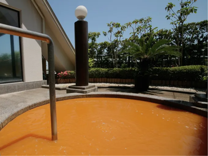 A round stone open-air bath filled with reddish-brown Kinsen hot spring water surrounded by natural landscaping.
